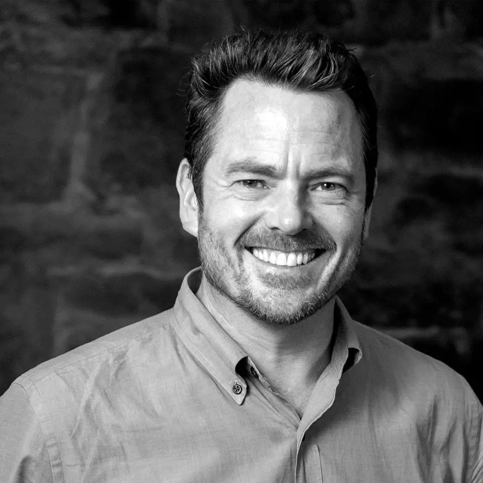 Screenshot A man smiling for the camera in front of a brick wall.